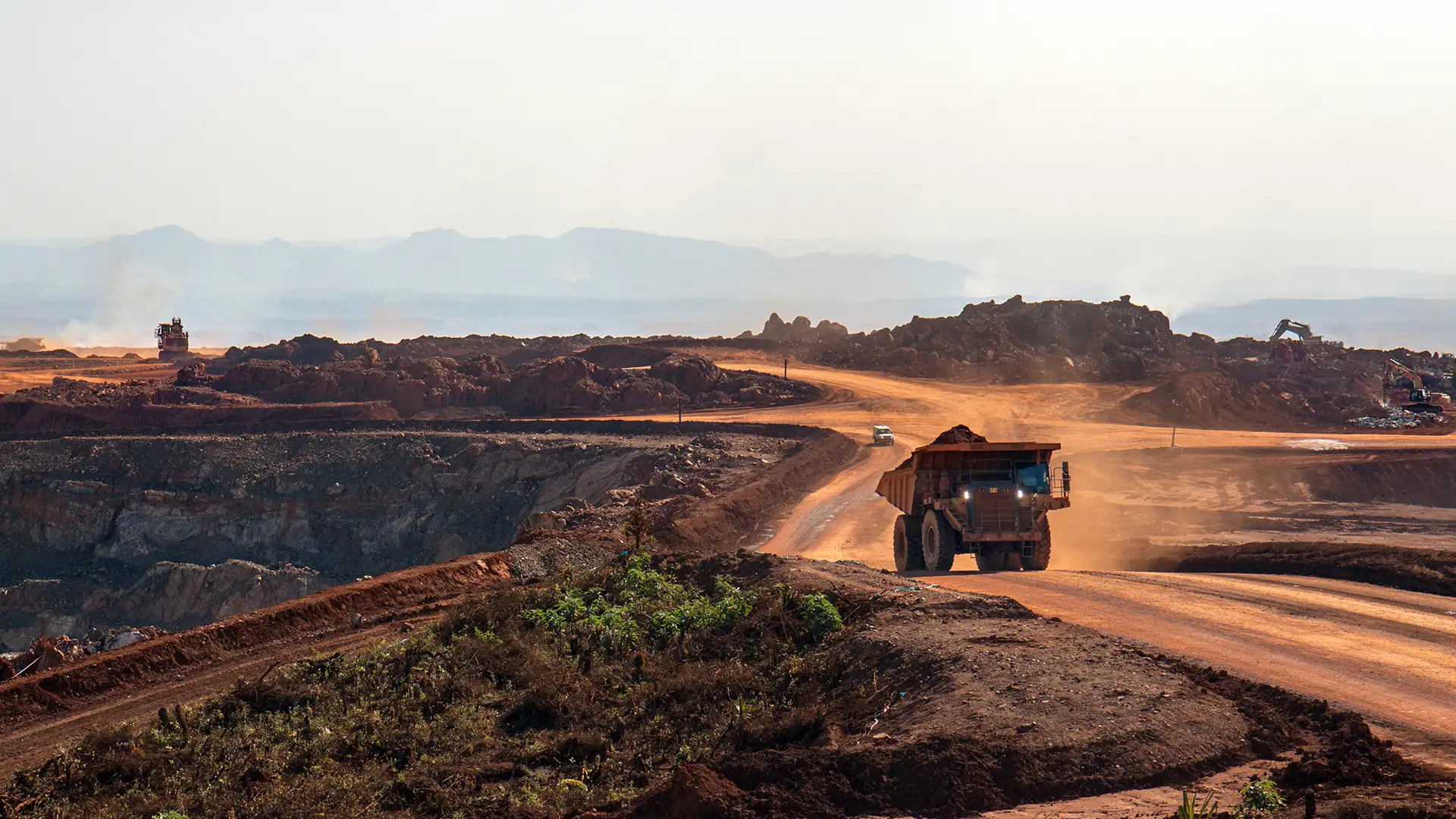 Large haul truck carrying mined earth along a dusty road in an open-pit mining site with surrounding excavation equipment.