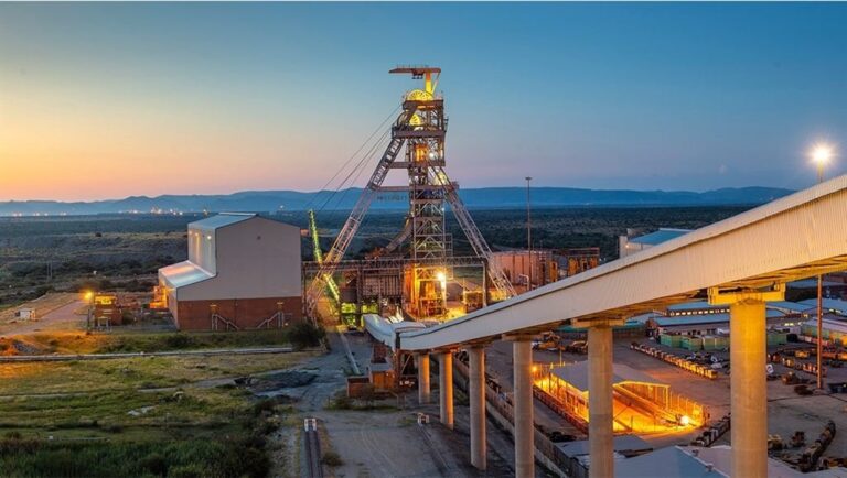 Mining facility at sunset with a tall lattice crane, conveyor belt, and lit structures over a desert landscape