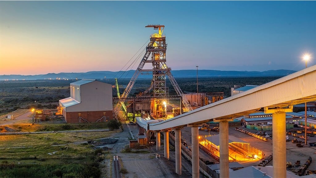Mining facility at sunset with a tall lattice crane, conveyor belt, and lit structures over a desert landscape